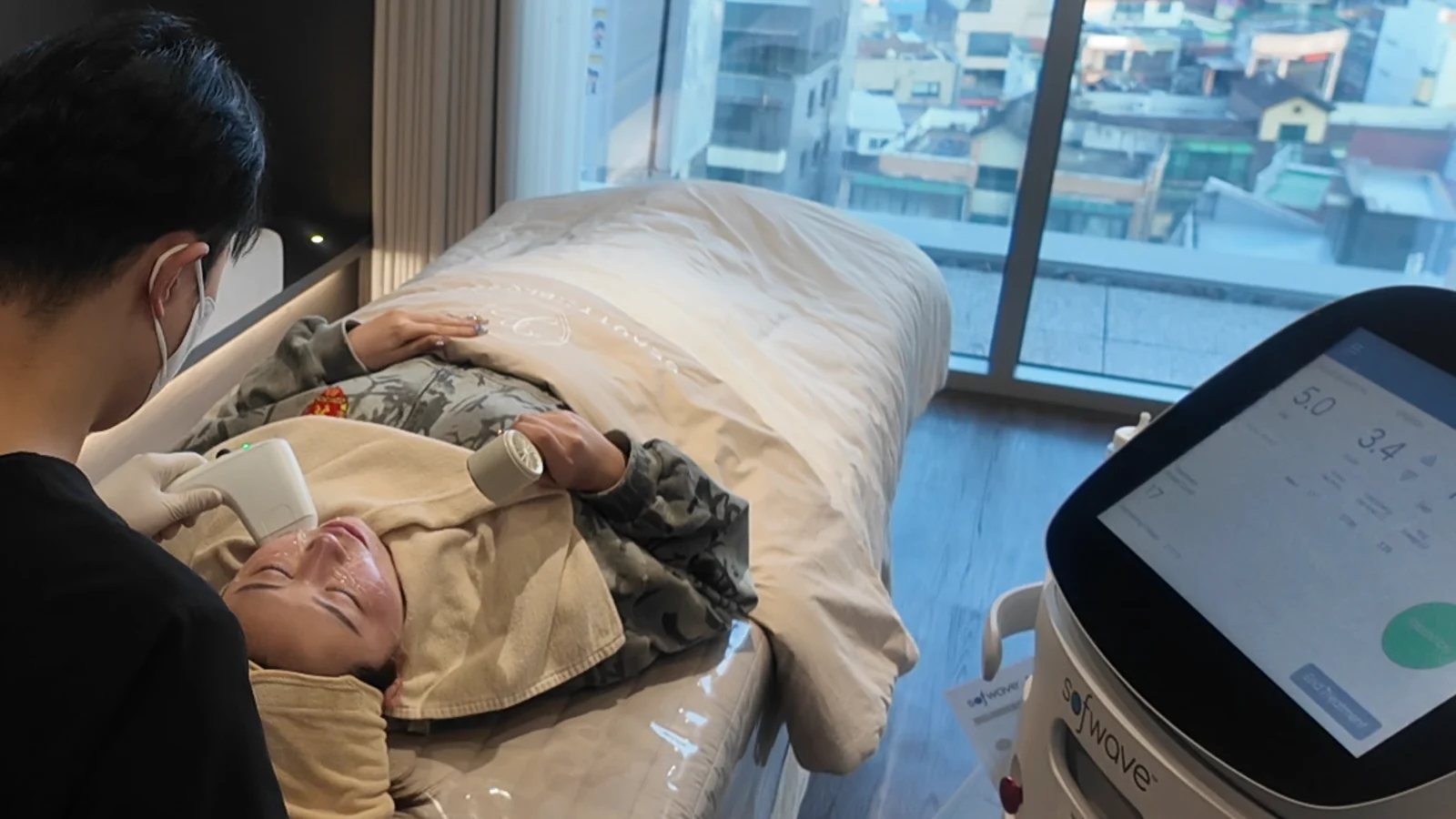 A person on a treatment bed beside a console while another person is seen from behind in a clinic room.