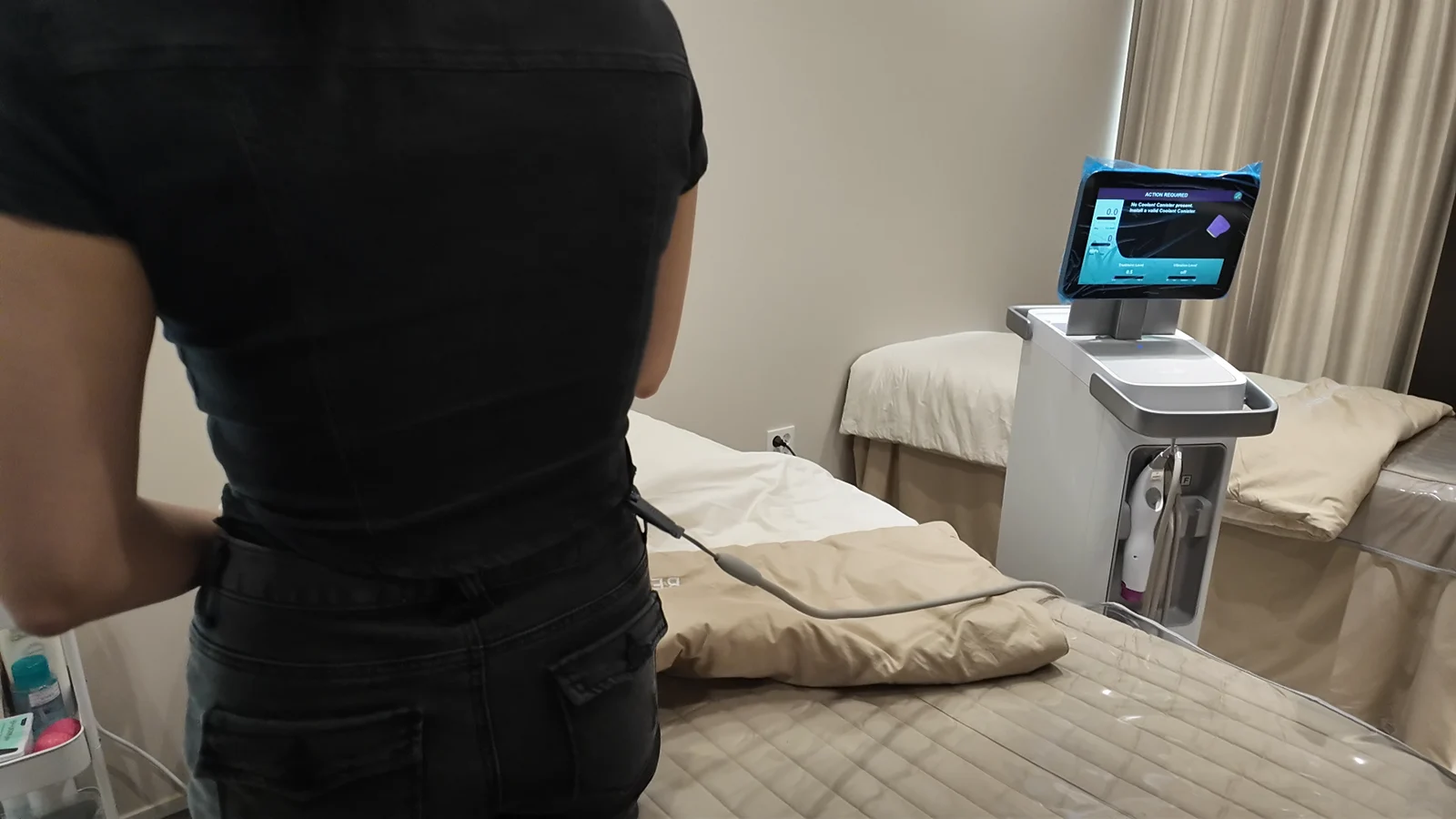 A person in black clothing stands beside treatment equipment in a clinic room.