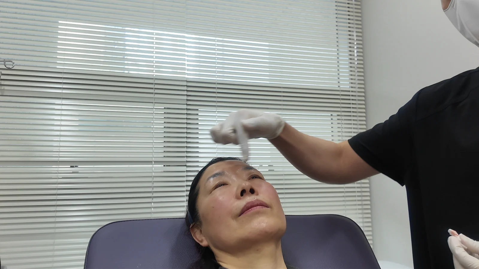 A woman wearing a black hairband lies on a clinic bed with a white tool near her forehead.