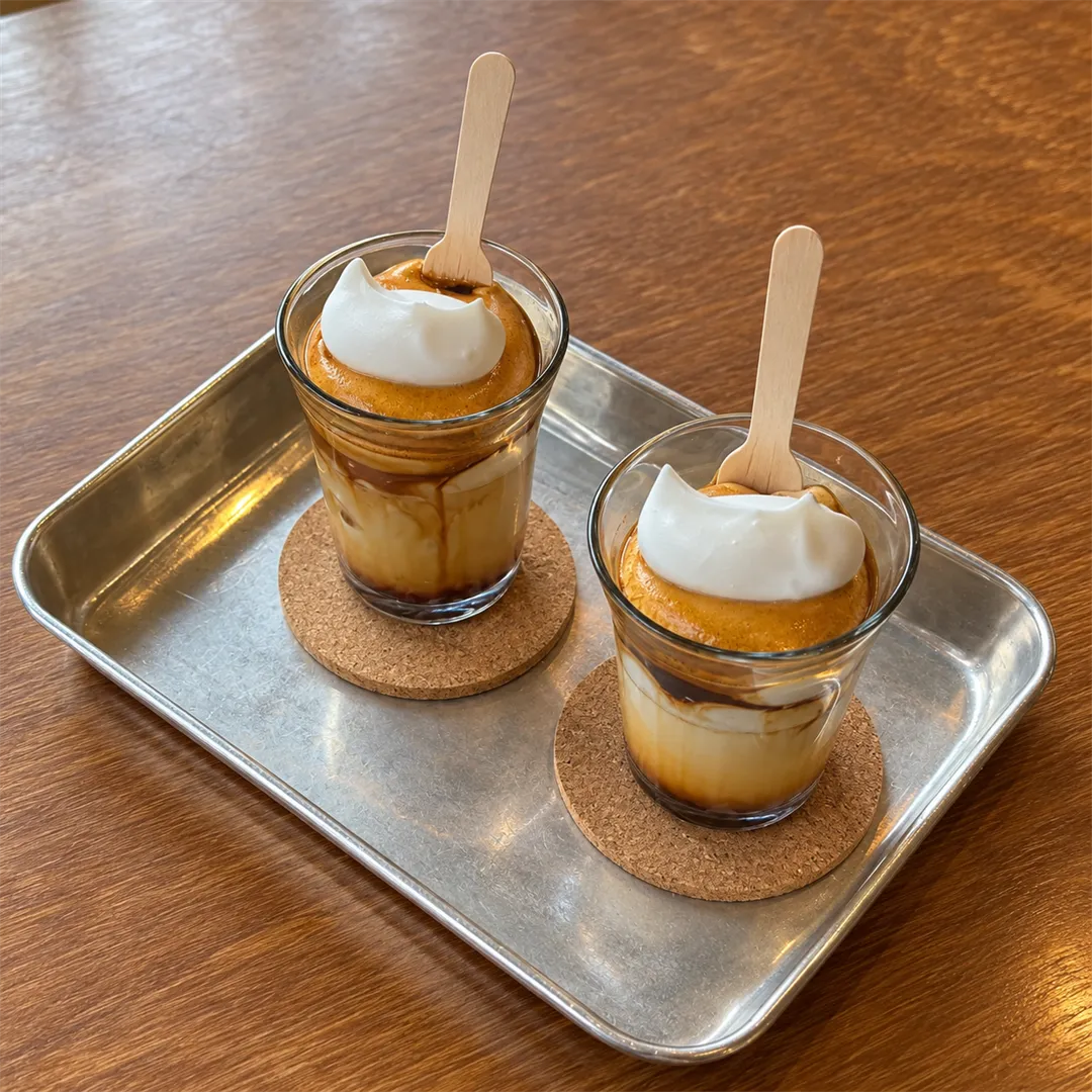 Two clear glasses of coffee topped with white cream on a metal tray with wooden spoons and cork coasters