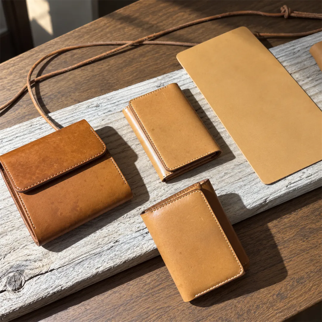 Three tan half wallets and a large flat tan leather panel on a light grey board with a thin leather strap