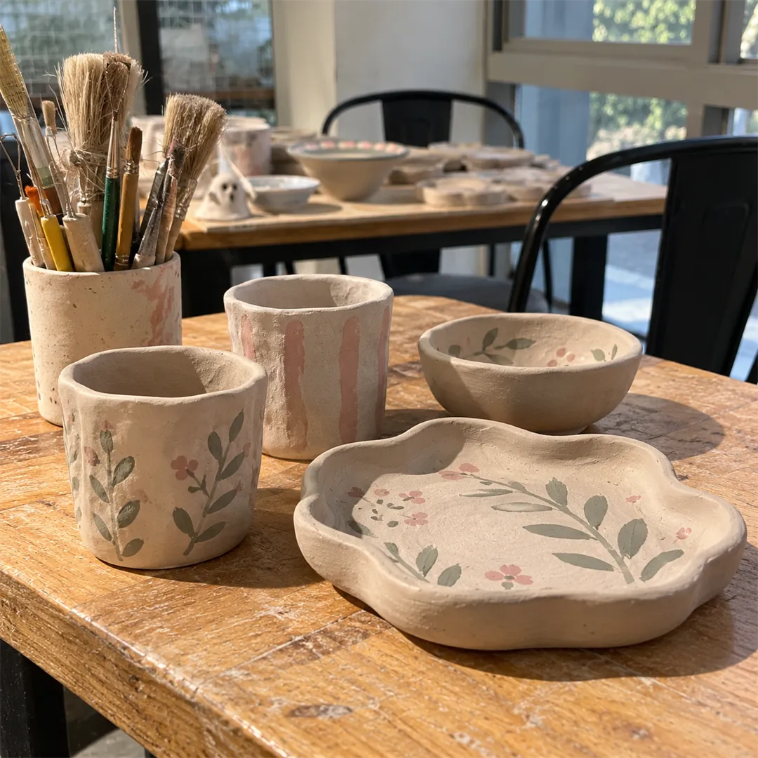 Small unfinished clay cups and bowls and a painted plate on a wooden table