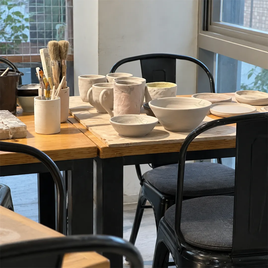 Wooden work table by a window with black chairs, a brush holder, cups, bowls and plates
