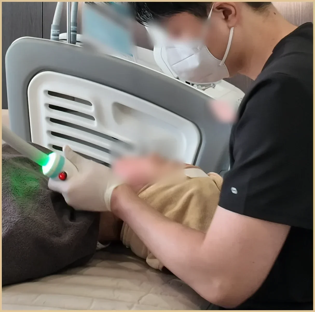 A masked clinician in dark scrubs holding a handpiece with a green light near a person lying down, with a treatment arm and device behind; eyes, face, a paper sheet and a note are blurred.