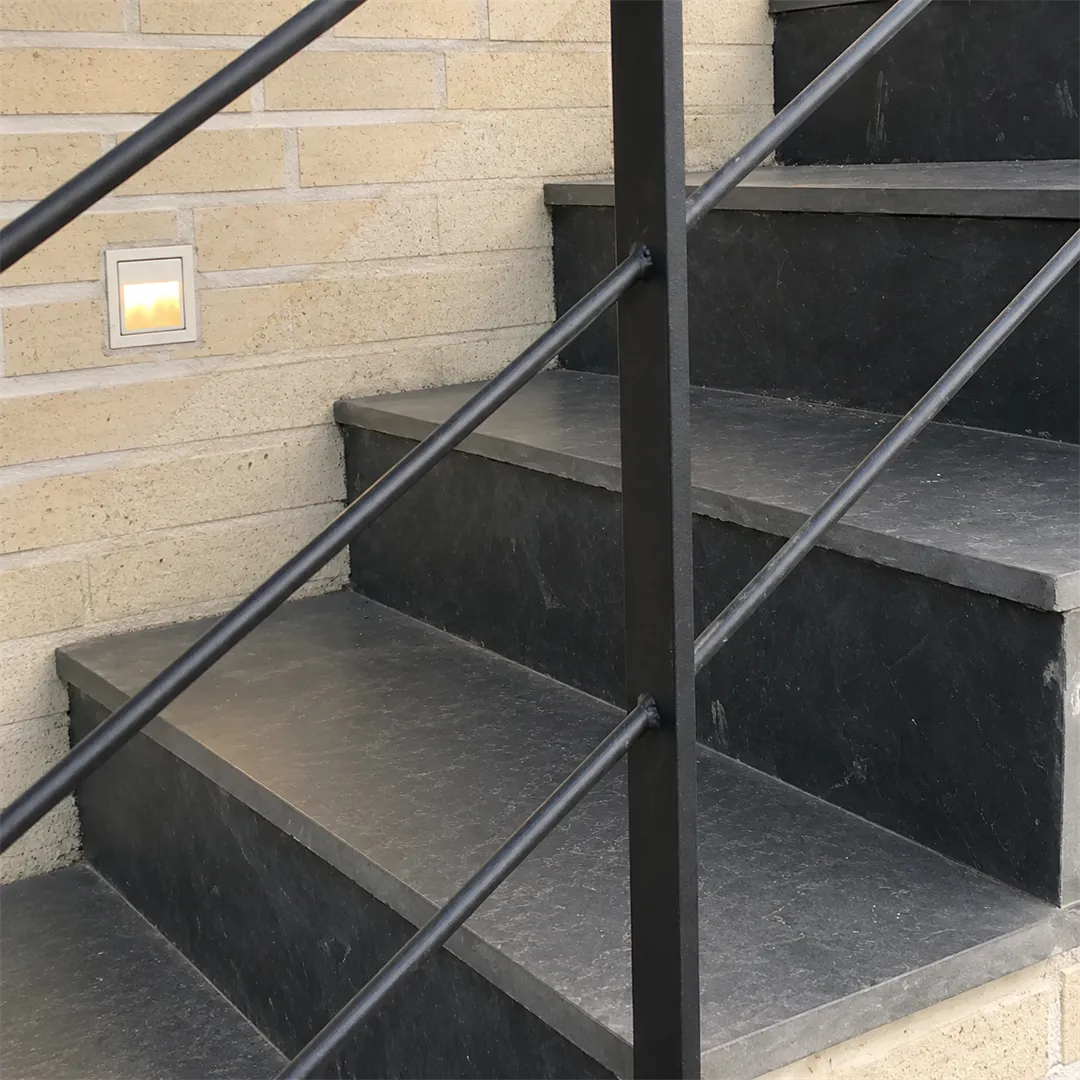 Black stone stairs beside a cream brick wall with a black metal railing and small warm wall lights