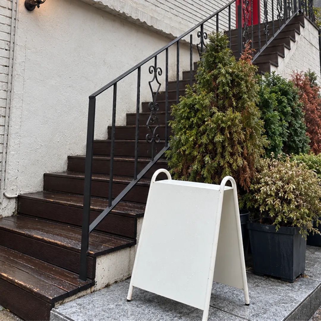 Dark wooden stairs with a black railing along a white brick wall with a potted plant and a blank white sign