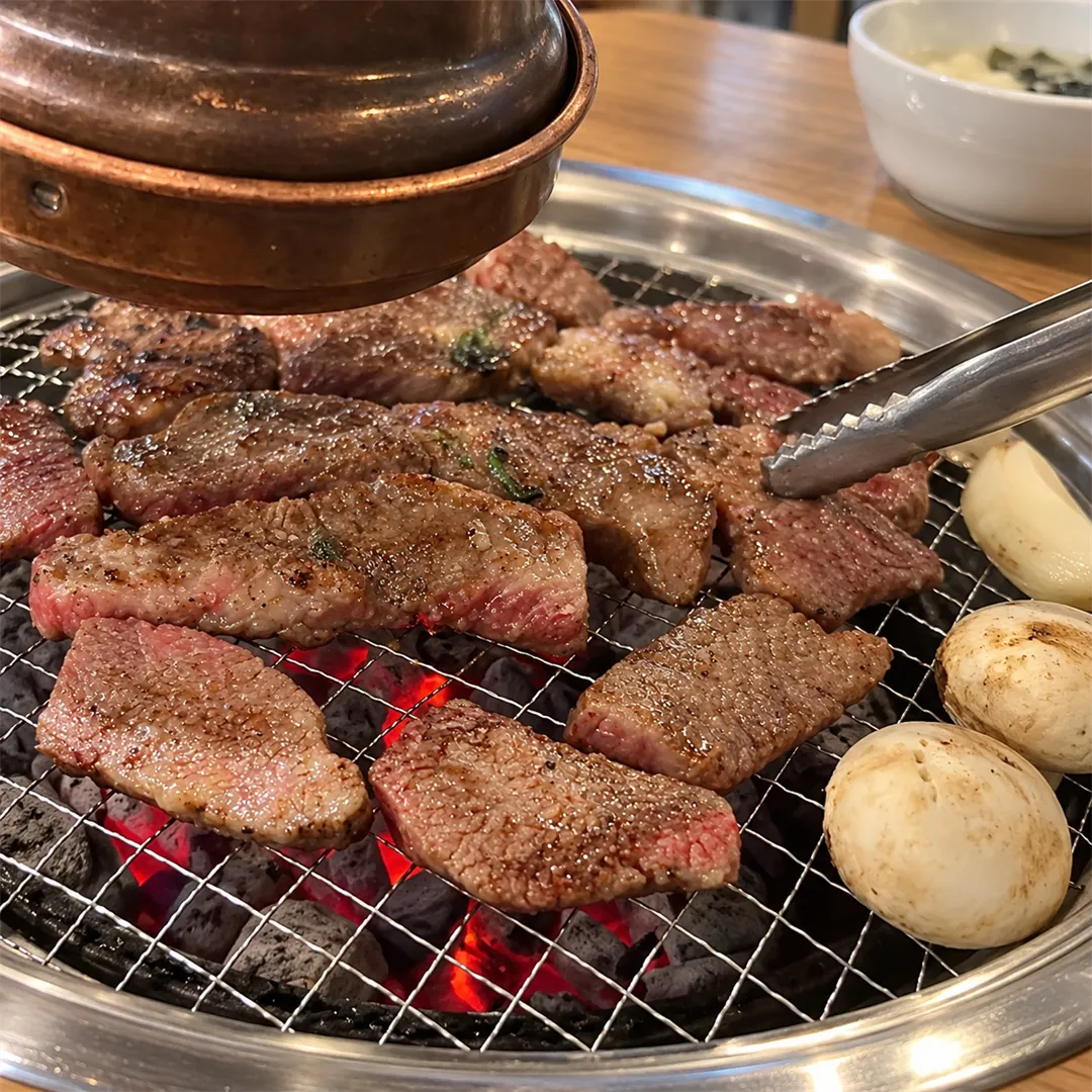 เนื้อวัวกำลังสุกบนตะแกรงถ่านใต้ท่อดูดควัน มีคีมและเห็ดวางข้าง
