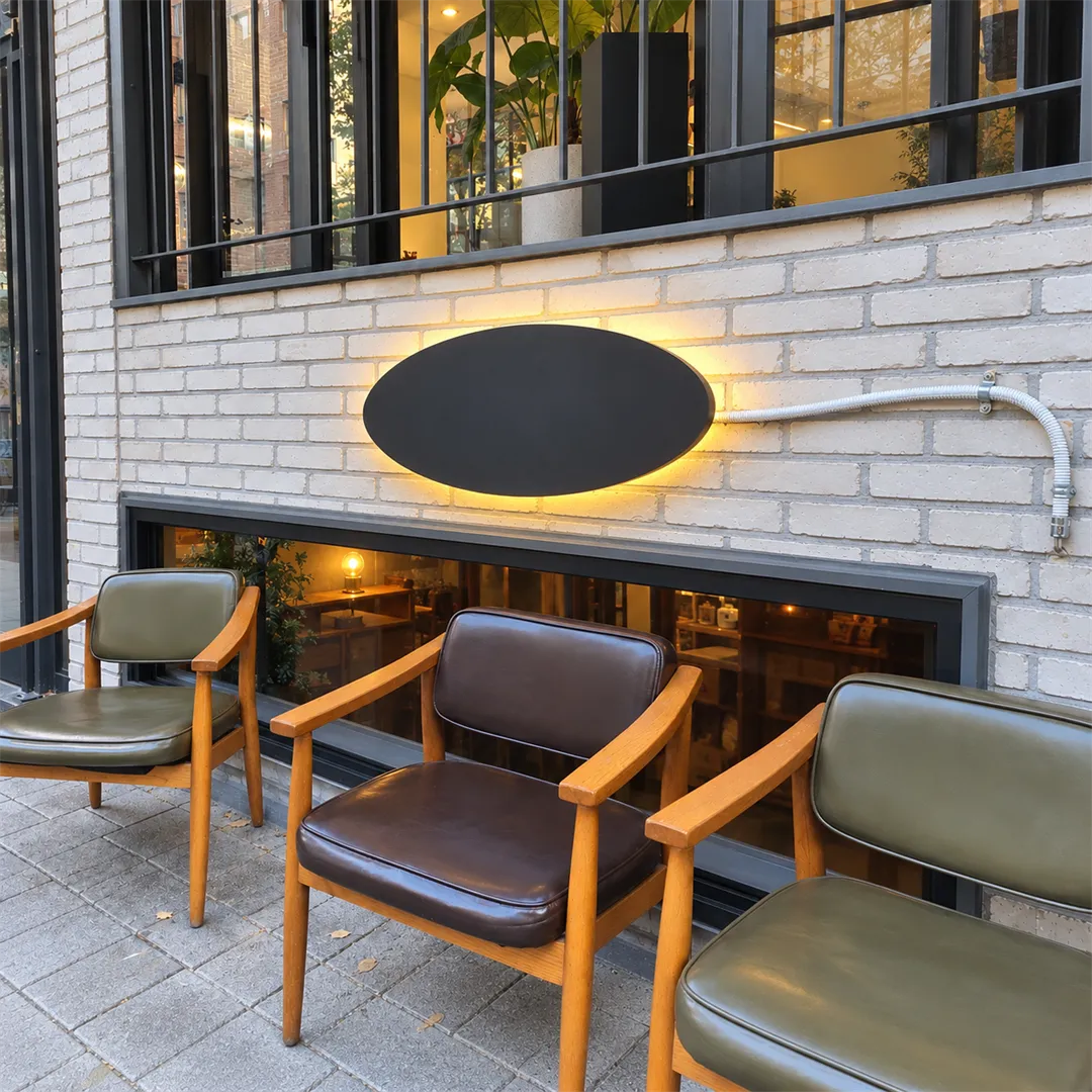White brick building with long windows, a dark oval sign and chairs outside