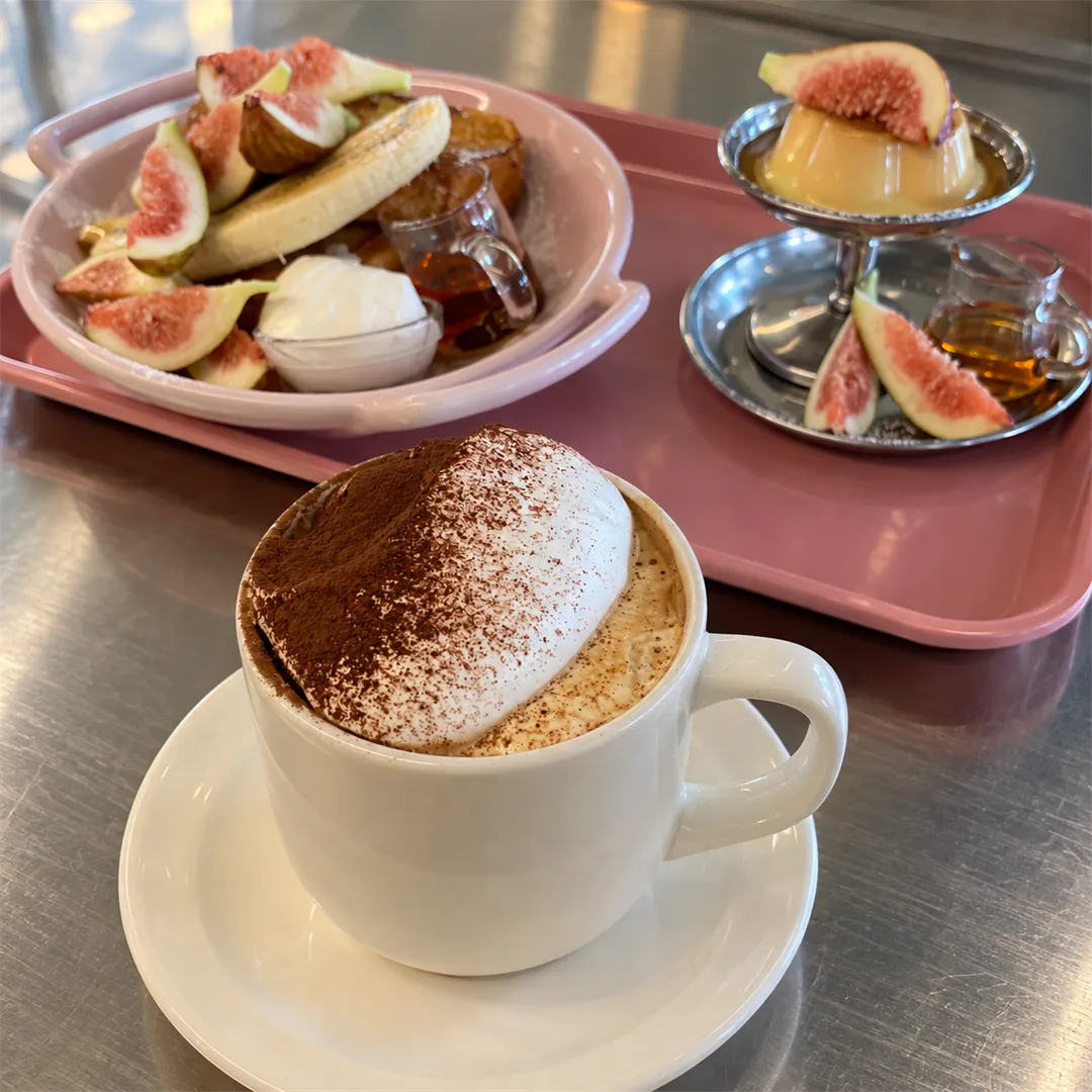 แก้วมักกาแฟที่มีครีมด้านบนวางบนจานรอง ด้านหลังเป็นกระทะสีชมพูและของหวานท็อปมะเดื่อ