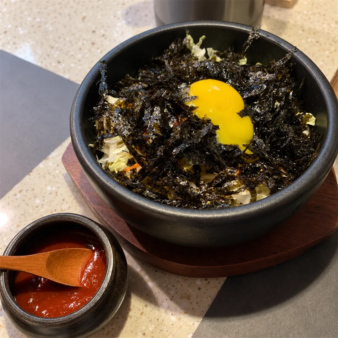 ชามหินสีดำมีข้าว ผักซอย สาหร่ายโรย และไข่แดงตรงกลาง ข้างถ้วยซอสแดงเล็ก