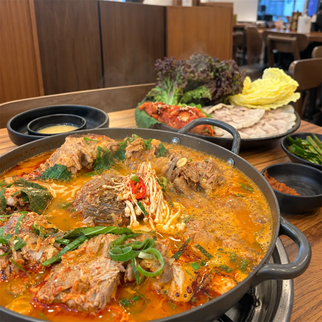 หม้อต้มกระดูกหมูน้ำซุปสีแดง มีเห็ดเข็มทองและต้นหอม ด้านหลังมีจานหมูต้มโปซัมกับกิมจิ