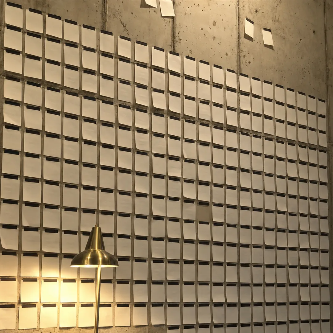 Concrete wall densely covered in small white paper notes with a brass cone floor lamp glowing below