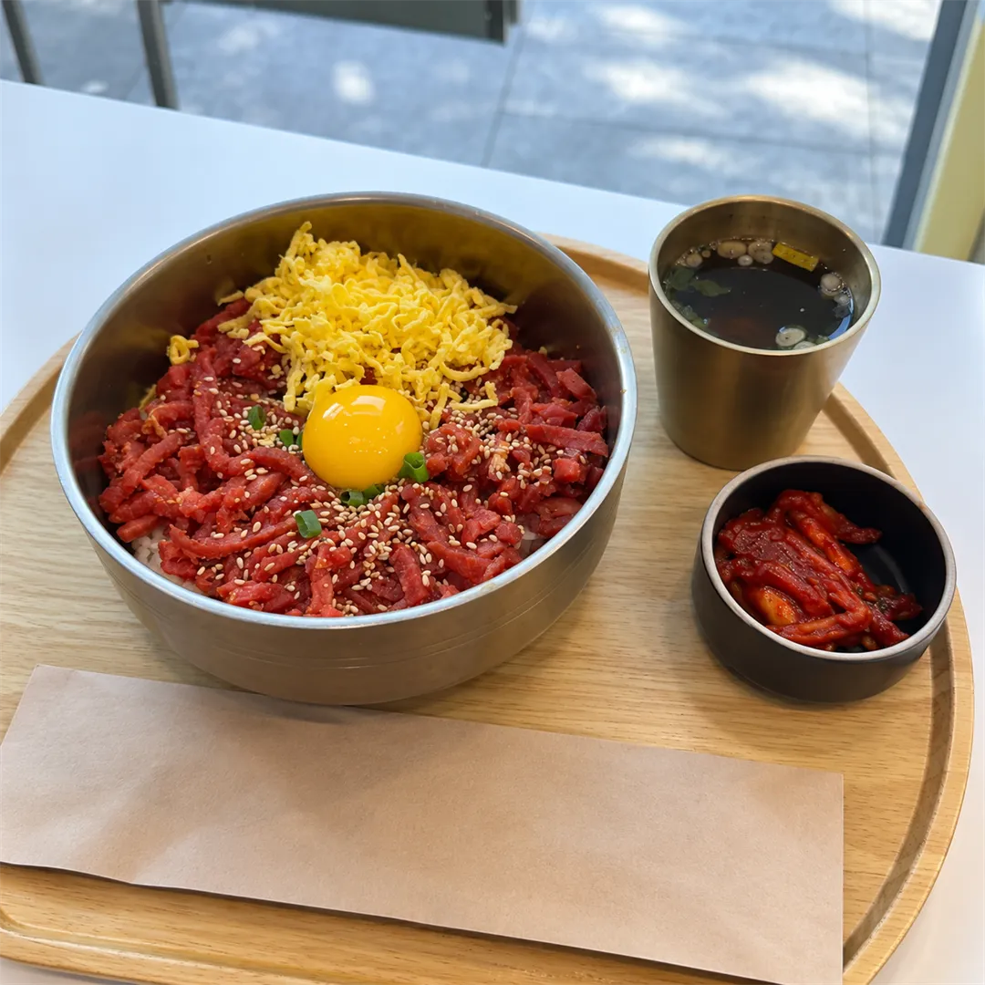Beef tartare rice bowl with an egg yolk, egg strips and sesame, with soup and kimchi on a wooden tray
