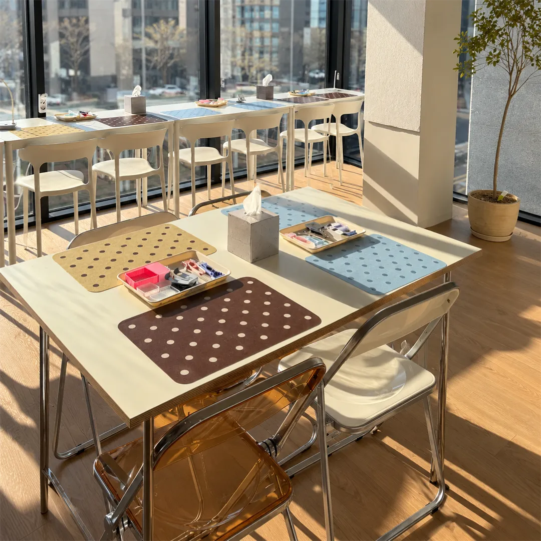 Sunlit work table with polka-dot mats, trays of beads and tweezers, and folding chairs by a window