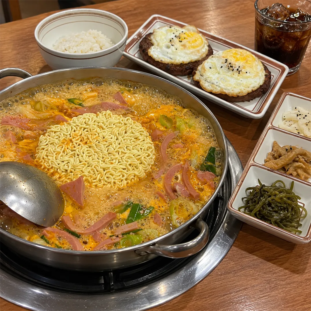 หม้อบูแดจิเกที่กำลังเดือดและมีบะหมี่ ด้านหลังมีจานแพตตี้สองแผ่นที่มีไข่ดาว ข้าว โค้ก และเครื่องเคียงสามช่อง