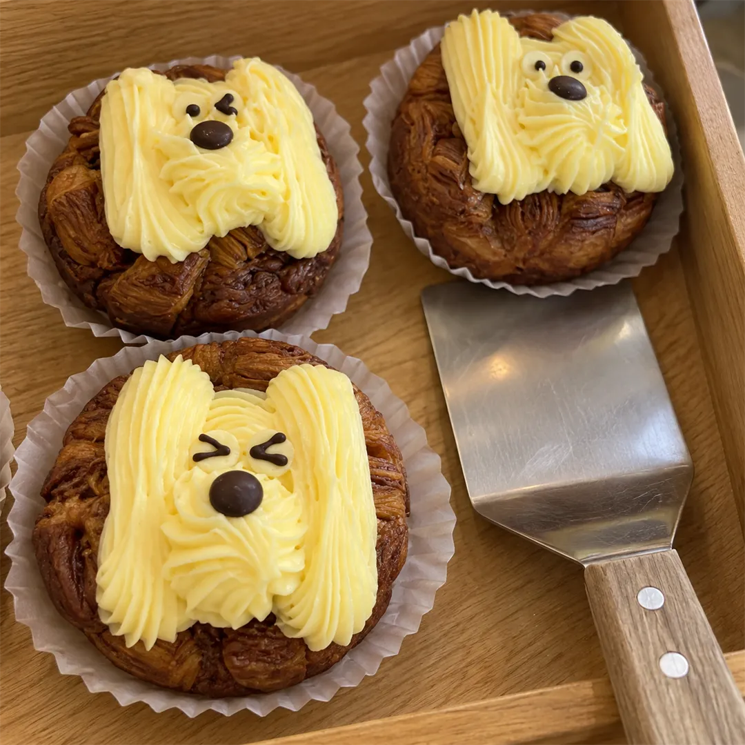 Three brown pastries decorated with yellow cream puppy faces in white paper cases on a wooden tray