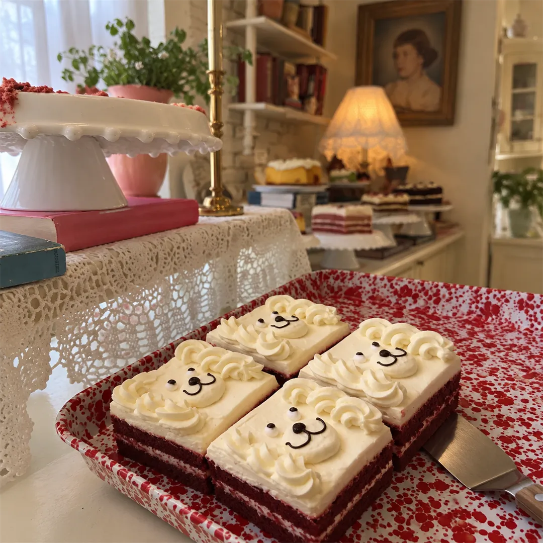 Four red velvet cake slices topped with cream faces on a red speckled tray, with a lace stand behind