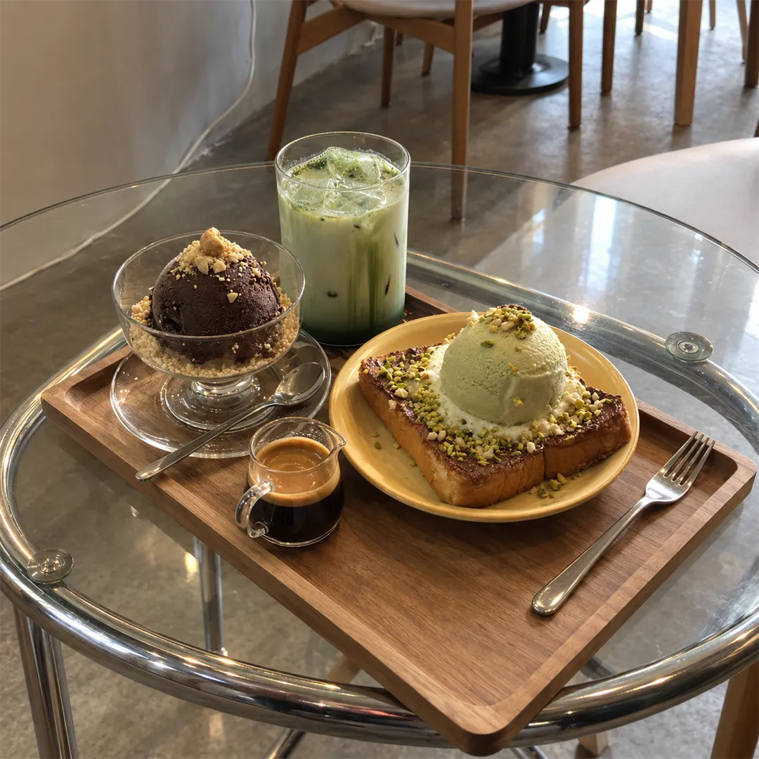 Wooden tray with a thick toast topped with a green scoop and chopped nuts, a glass of dark brown scoop dessert, a green iced drink and a small espresso jug