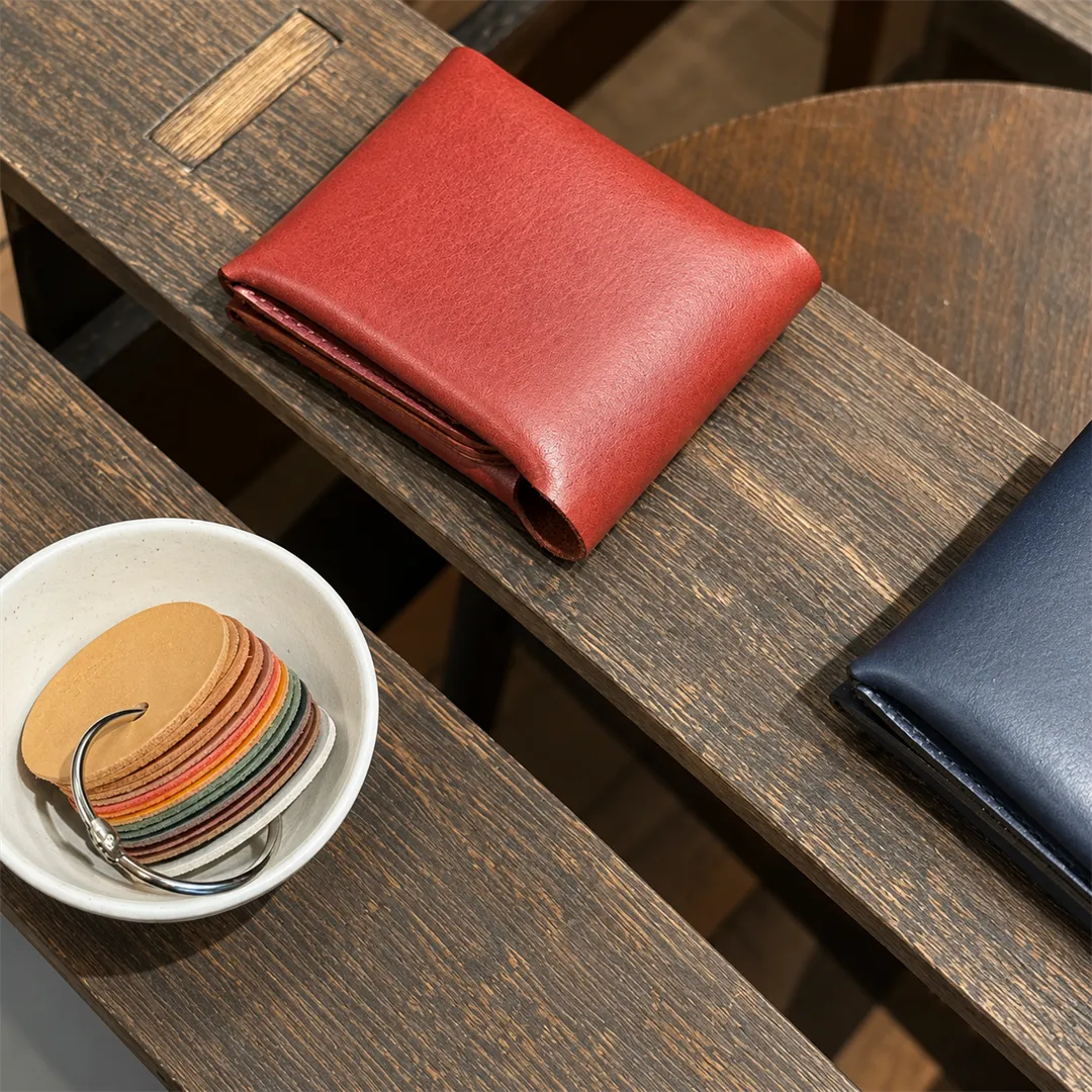 Red and navy leather wallets on a dark wooden slat and a white bowl of leather color samples strung on a silver ring