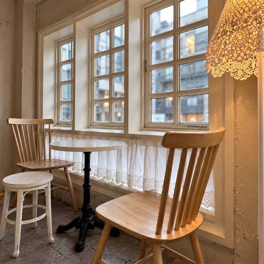 Window seat with pleated white curtains, a small round white table, two wooden chairs and a lace-shaded lamp