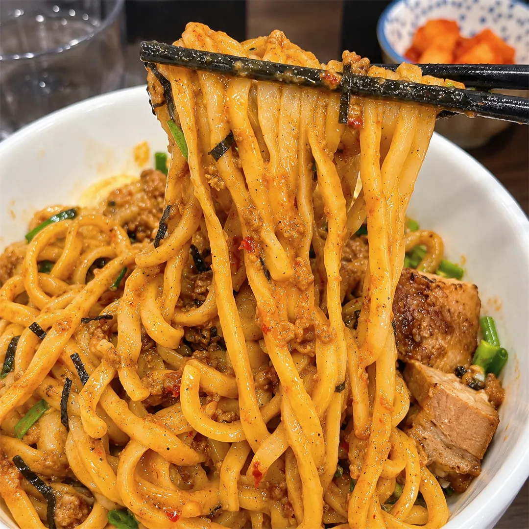 ตะเกียบดำยกเส้นบะหมี่หนาขึ้นจากชาม ด้านล่างเป็นเส้นเคลือบซอสและเนื้อสับ