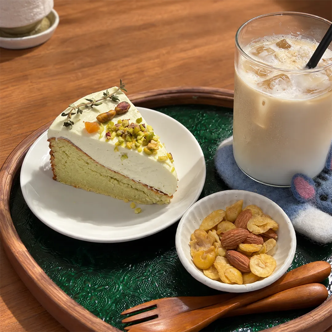 Cream cake with nut topping, a latte and a plate of nuts and cereal on a green round tray