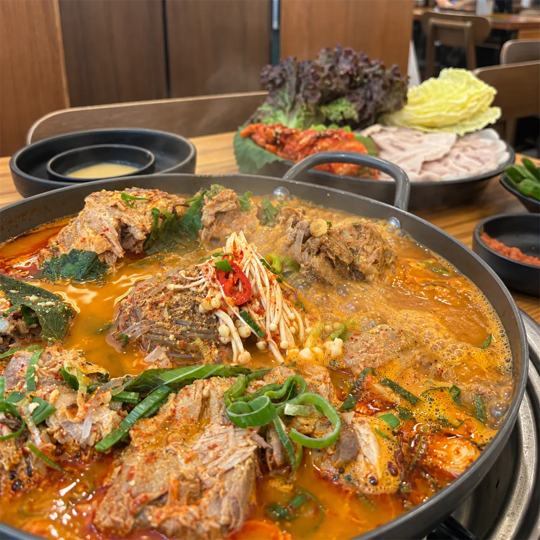ภาพใกล้ของหม้อต้มกระดูกหมู น้ำซุปแดงและต้นหอม มีจานโปซัมอยู่ด้านหลัง