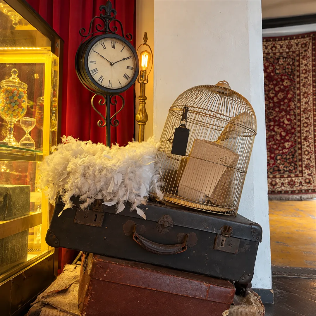 Decorative corner with a Roman numeral clock, a gold birdcage, red curtains, feather props and an old suitcase